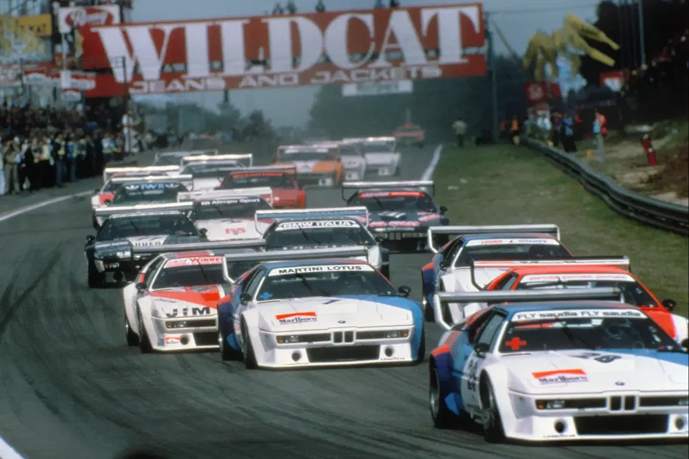 Course de voitures BMW M1 sur un circuit, foule en arrière-plan sous une bannière Wildcat.