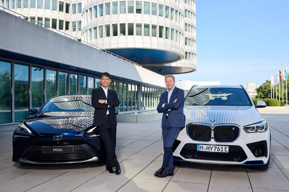 Toyota Mirai en BMW X5 voor een modern gebouw, vertegenwoordigers in gesprek.
