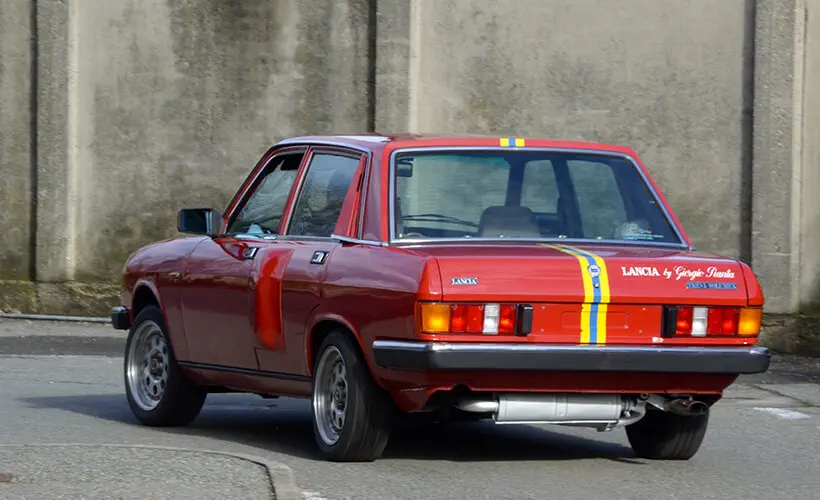 Lancia rouge avec bande jaune, en mouvement sur route. Modèle vintage à carrosserie carrée.