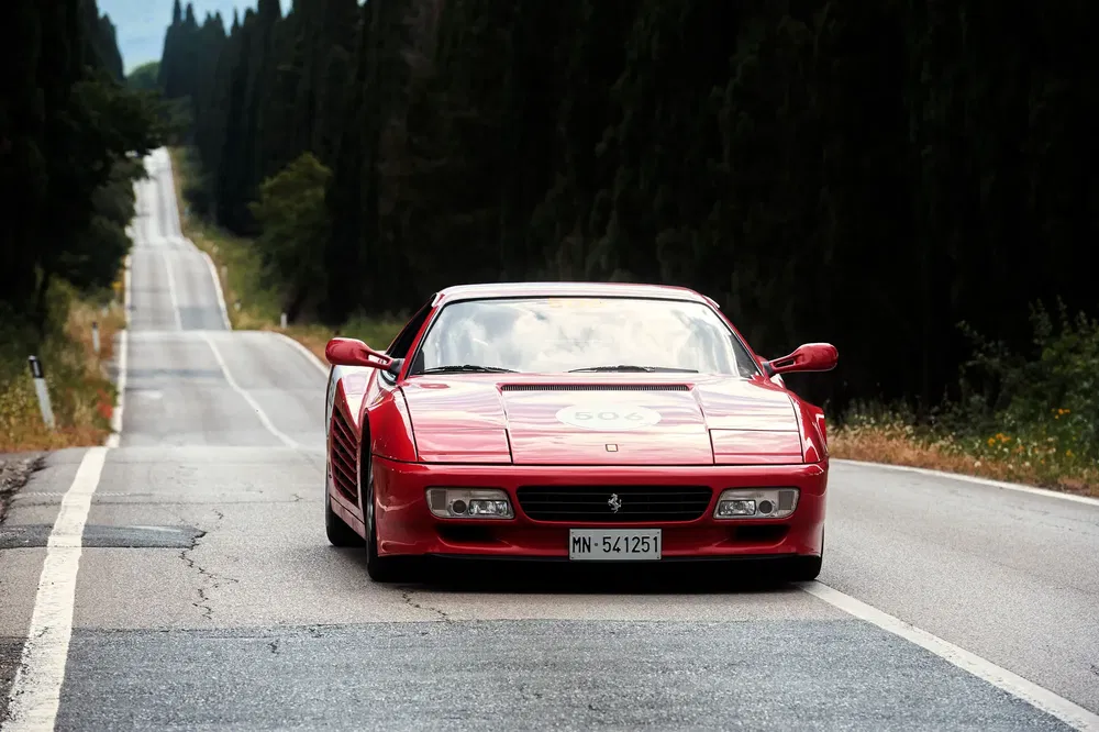 Ferrari Testarossa rouge sur une route bordée d'arbres, roulant en pleine campagne.