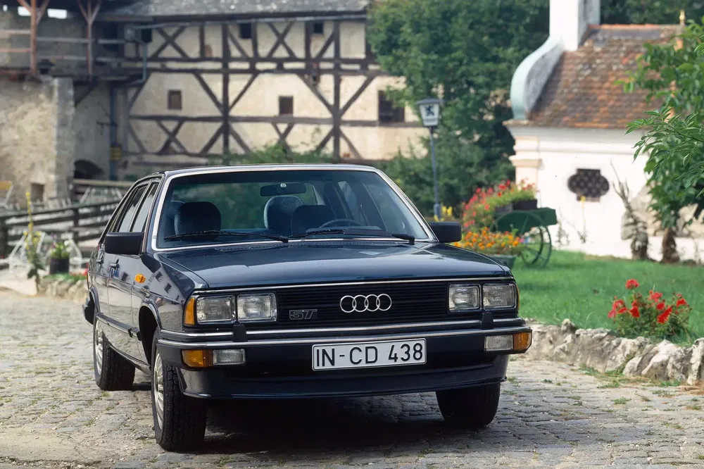 Audi 80 Avant noir dans une ruelle pavée, architecture médiévale en arrière-plan.
