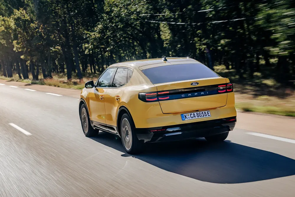 Ford Capri jaune roulant sur une route bordée d'arbres, vue arrière.