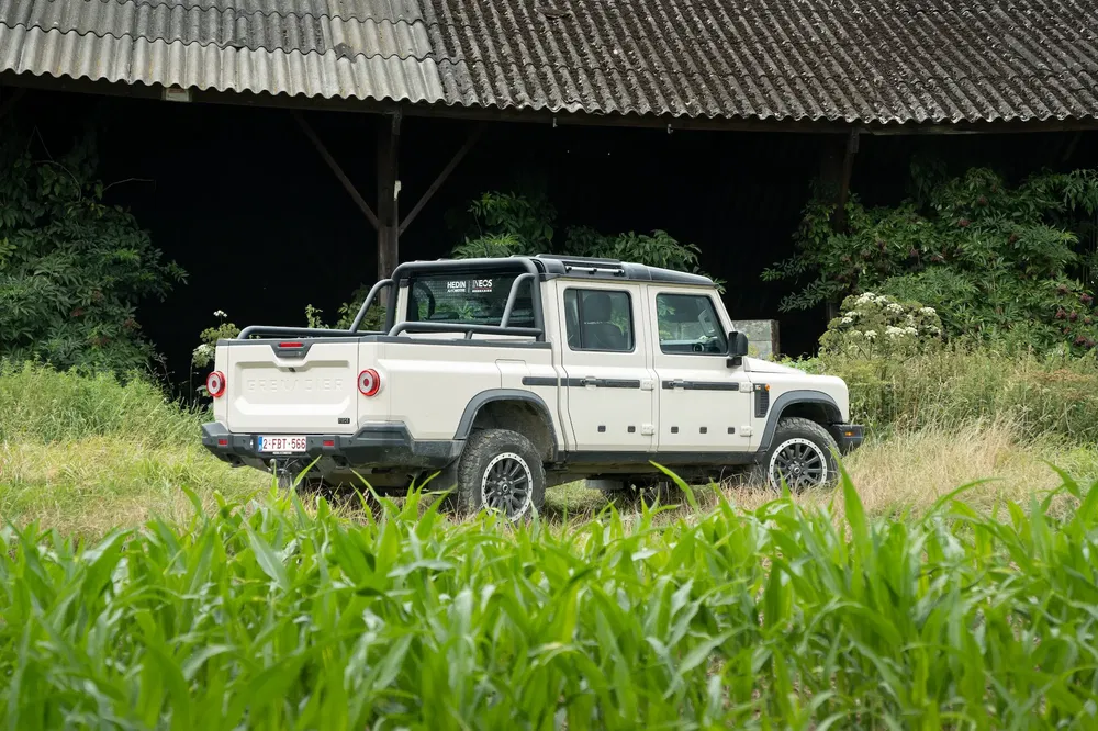 Witte Ineos Grenadier 4x4 truck in een veld naast een houten schuur.