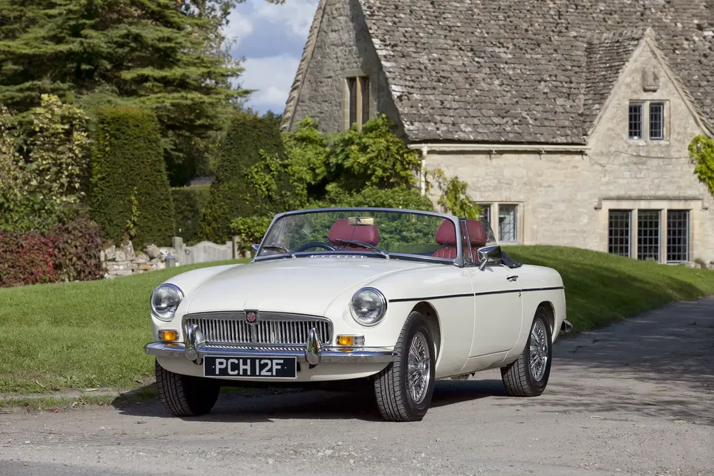 Voiture MG B blanche décapotable devant une maison en pierre pittoresque.