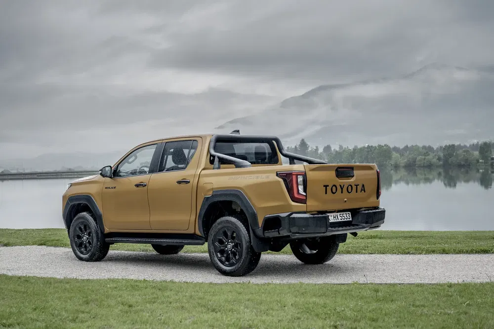 Toyota Hilux doré devant un lac sous un ciel nuageux.