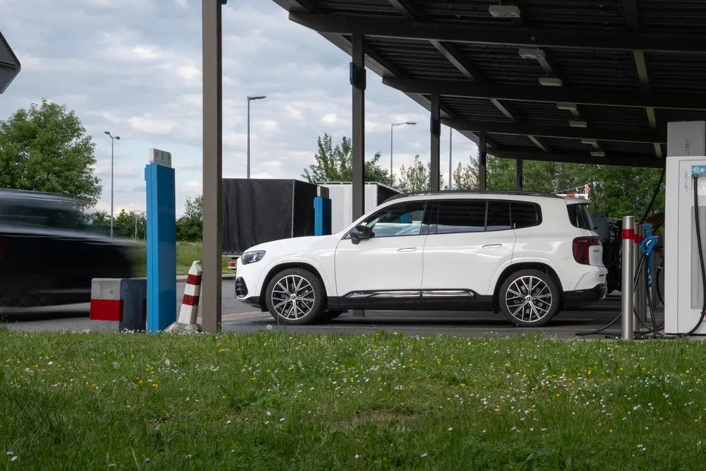 SUV blanc stationné à une station de recharge électrique, ciel nuageux en arrière-plan.