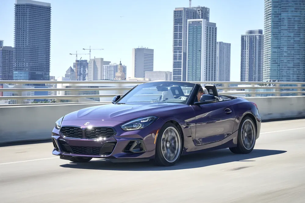 Paarse BMW Z4 cabriolet rijdt door stad met skyline op de achtergrond.