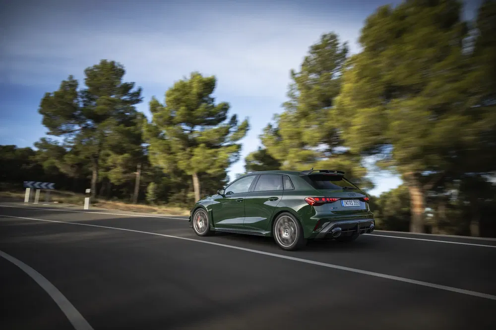 Audi RS3 verte roulant sur une route bordée d'arbres, vue arrière dynamique.