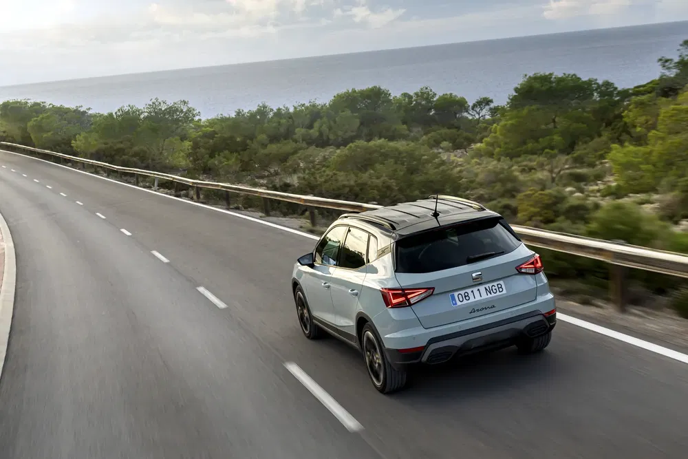 Grijze SEAT Arona SUV op kustweg met zee en bomen op de achtergrond.