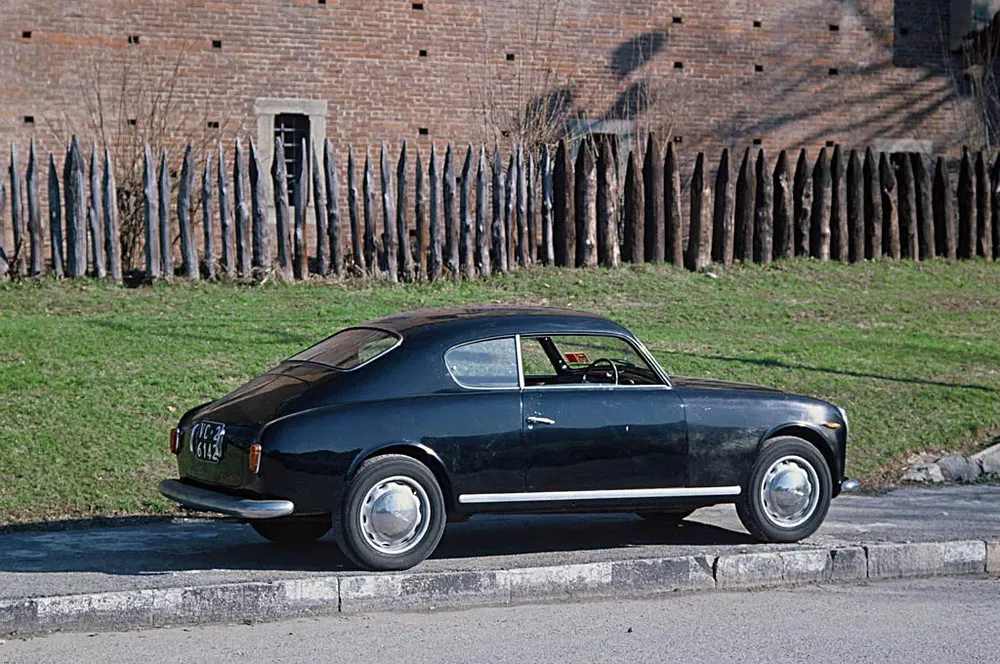 Alfa Romeo vintage noire garée devant une palissade en bois et un mur en brique.