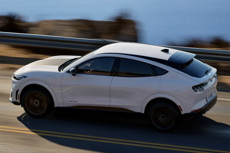Ford Mustang Mach-E blanche roulant sur une route côtière, vue en mouvement latéral.
