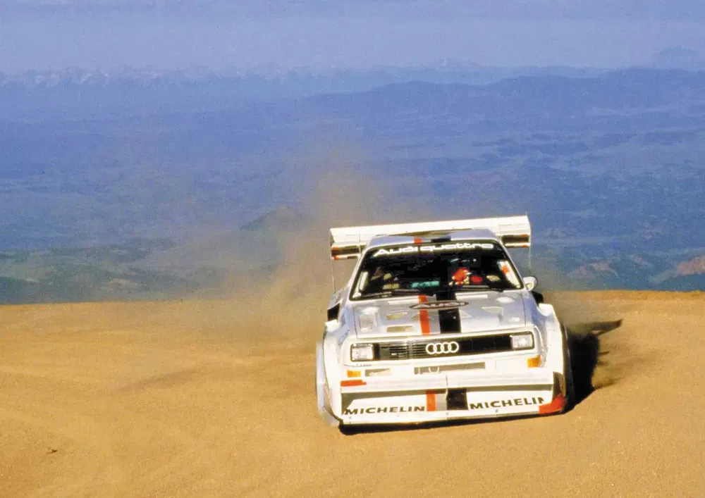 Audi Quattro blanche fonçant sur une piste de rallye poussiéreuse, montagnes en arrière-plan.