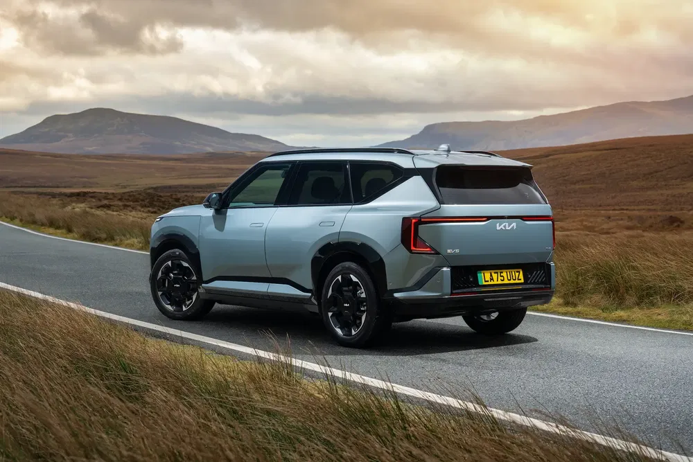 Kia EV9 SUV rijdt op een landelijke weg met bergen op de achtergrond.