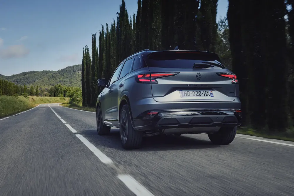 Renault Austral op een landelijke weg, dynamische achterzijde, omgeven door bomen en heuvels.