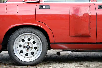 Voiture rouge avec un panneau de porte endommagé, jante blanche et échappement visible.