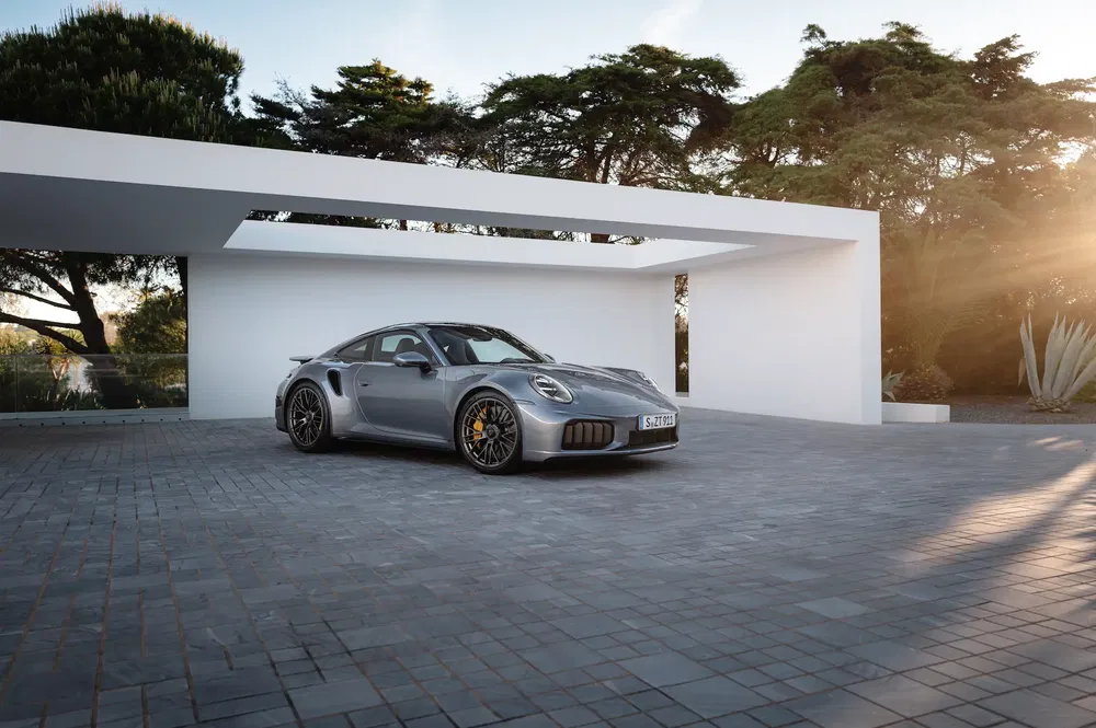 Porsche 911 Turbo S grise garée sous un auvent moderne, éclairée par le soleil couchant.