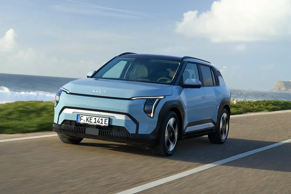 Kia EV9 roulant sur une route côtière avec vue sur l'océan, ciel partiellement nuageux.