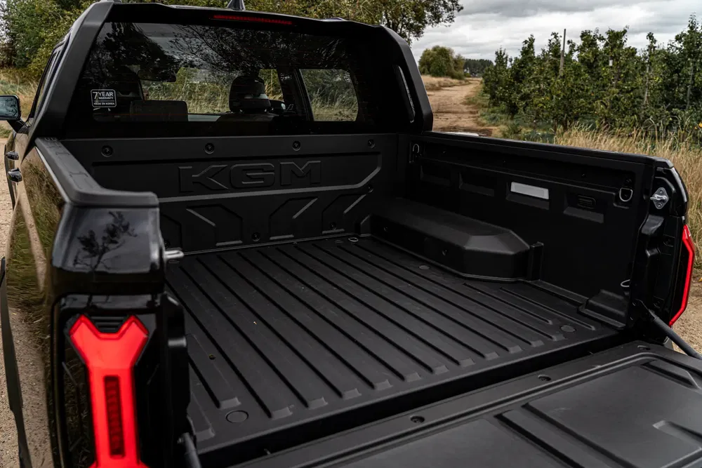 Benne de pick-up KGM avec hayon abaissé sur chemin de terre bordé d'arbustes.