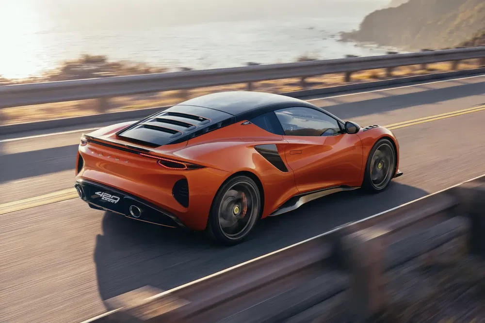 Lotus Emira orange roulant sur une route côtière ensoleillée, design aérodynamique élégant.