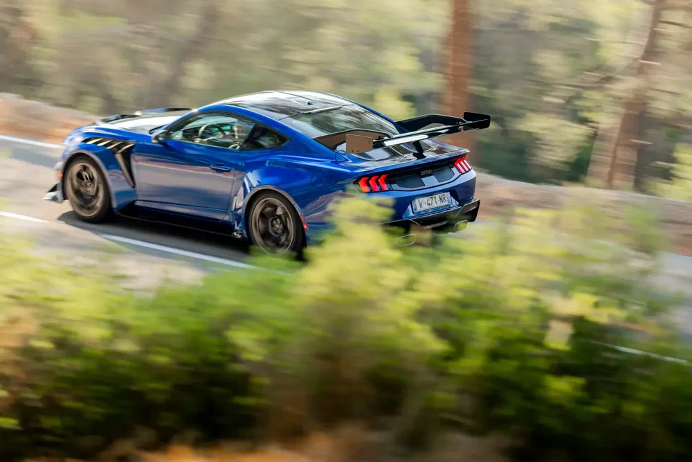 Voiture bleue sportive Ford Mustang GT3 roulant rapidement sur une route bordée d'arbres.