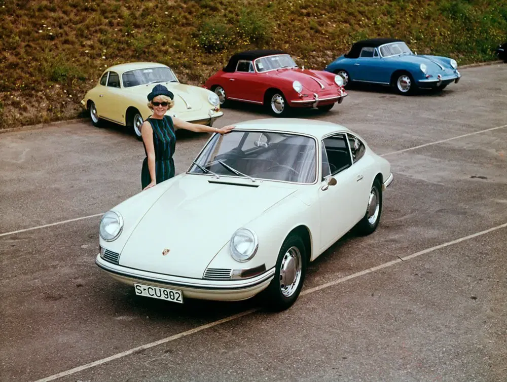Klassieke witte Porsche 911 met verschillende gekleurde modellen op de achtergrond op een parkeerplaats.
