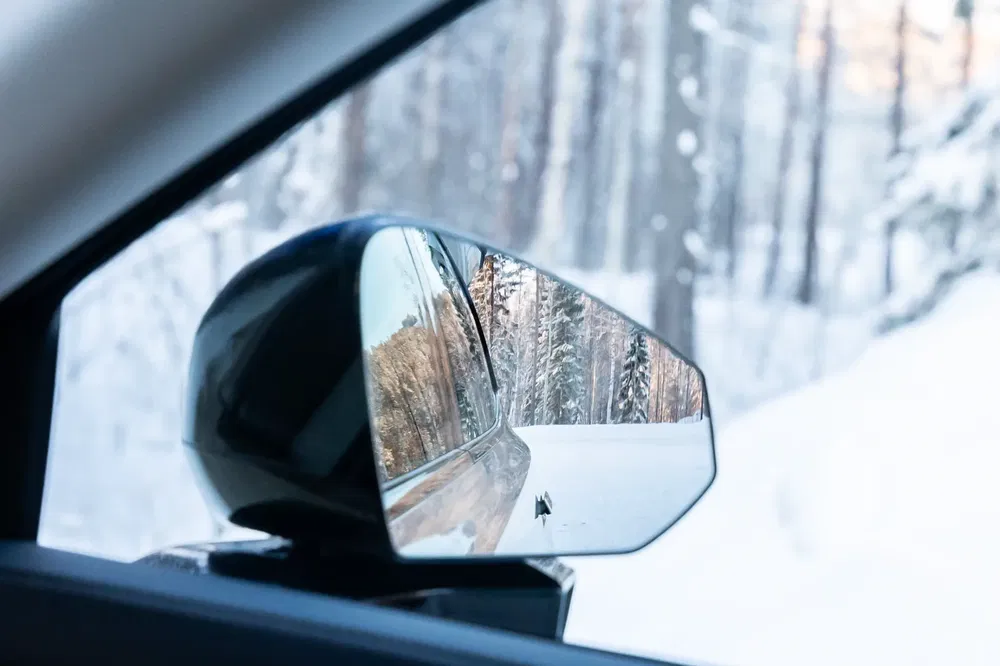 Rétroviseur de voiture sur route enneigée avec réflexion de la forêt hivernale.