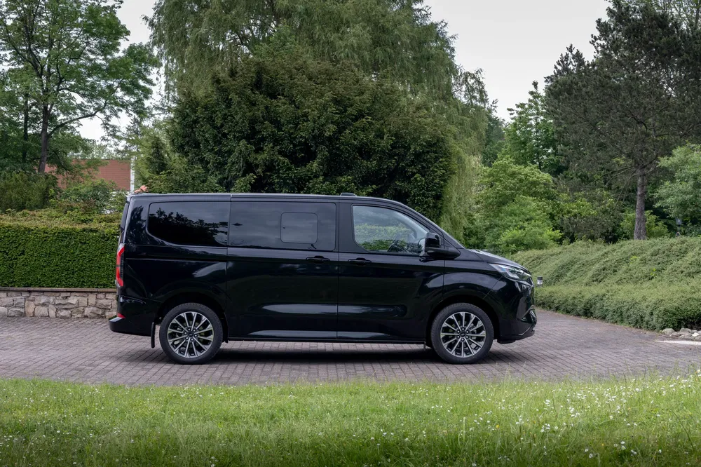 Fourgon Ford Custom noir garé sur une allée pavée entourée de verdure dense.