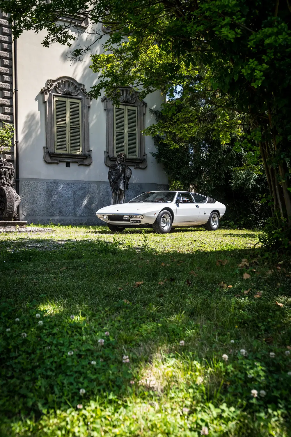 Witte Lamborghini Urraco voor een historische woning, omgeven door groen en beelden.