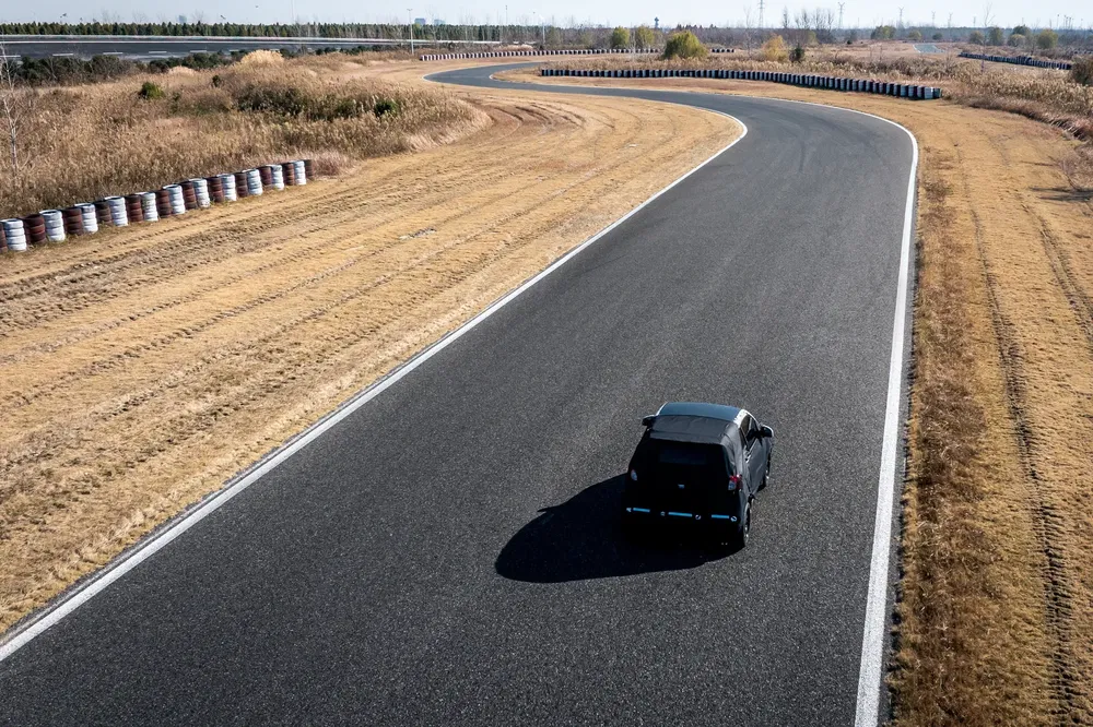 Voiture noire sur piste sinueuse, bordée d'herbes sèches et de pneus empilés.