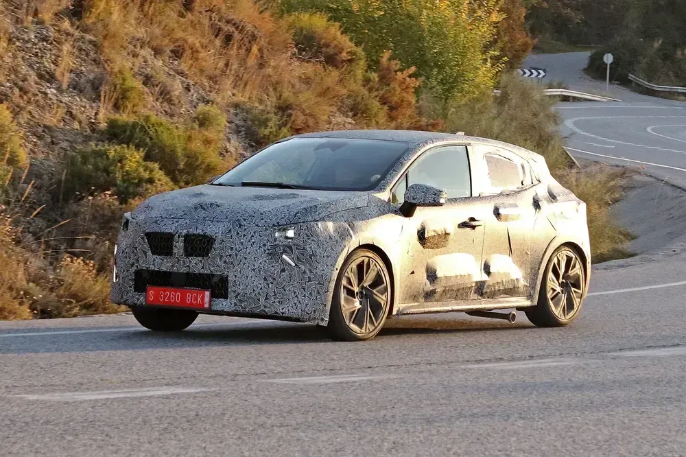 Voiture camouflée en test sur route sinueuse, entourée de végétation verte.