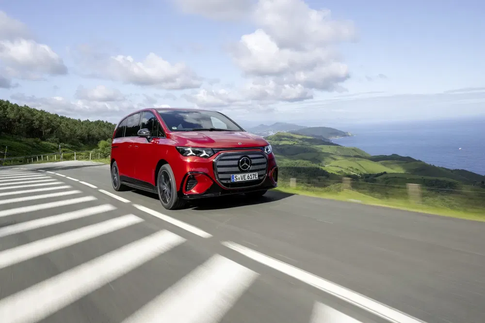 Mercedes Classe V rouge sur route côtière, paysage verdoyant en arrière-plan.