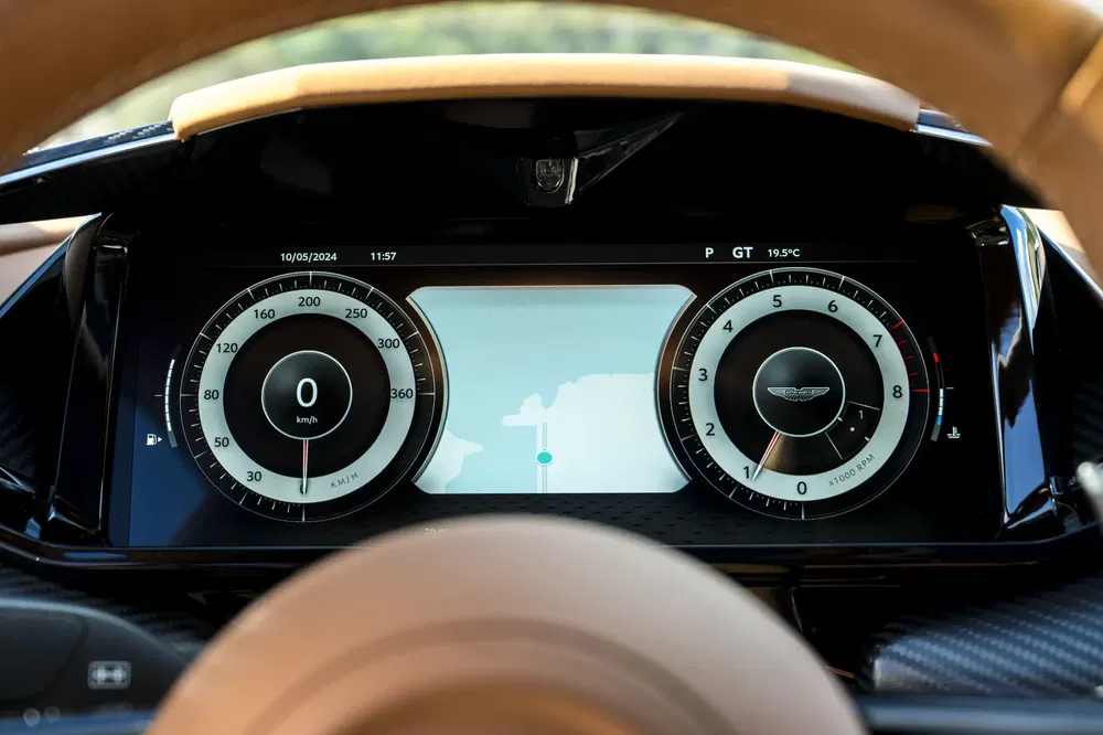 Tableau de bord numérique Aston Martin avec compteur de vitesse, navigation, et logo emblématique.