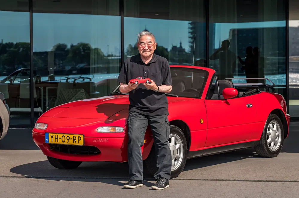 Man poseert met een rode Mazda MX-5 cabrio voor een modern gebouw.