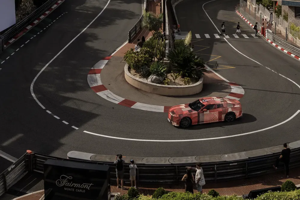 Voiture camouflée sur circuit Fairmont Monaco, virage serré, entourée de spectateurs.