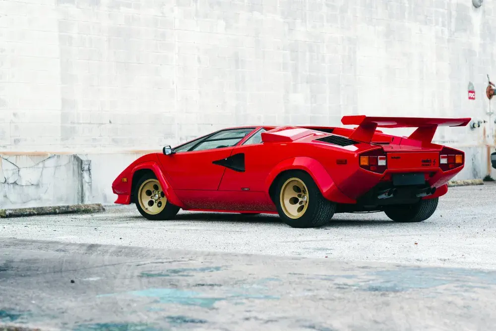 Rode Lamborghini Countach met achterspoiler en gouden velgen, geparkeerd op stedelijke achtergrond.