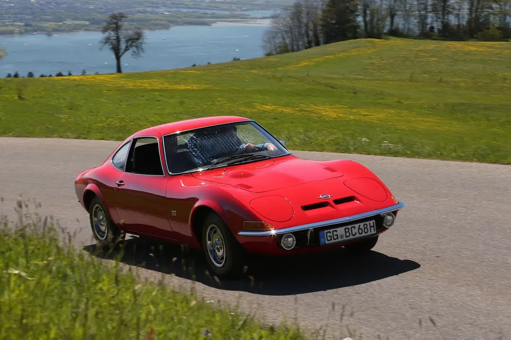Rode Opel GT op een schilderachtige weg met uitzicht op een meer in de zomer.