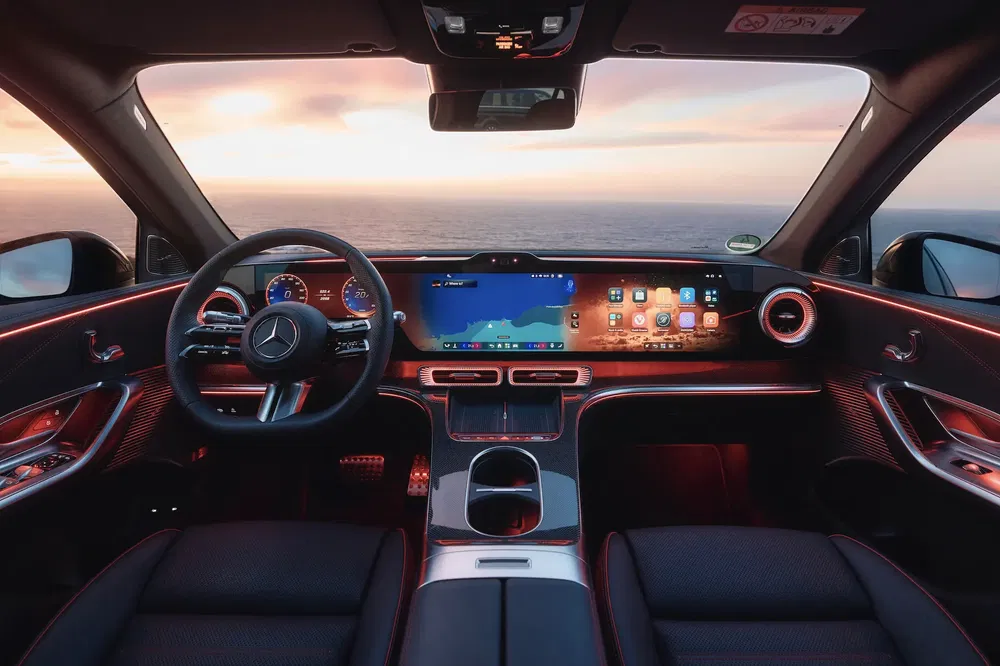 Interieur van een Mercedes met een digitaal dashboard en uitzicht op de oceaan bij zonsondergang.