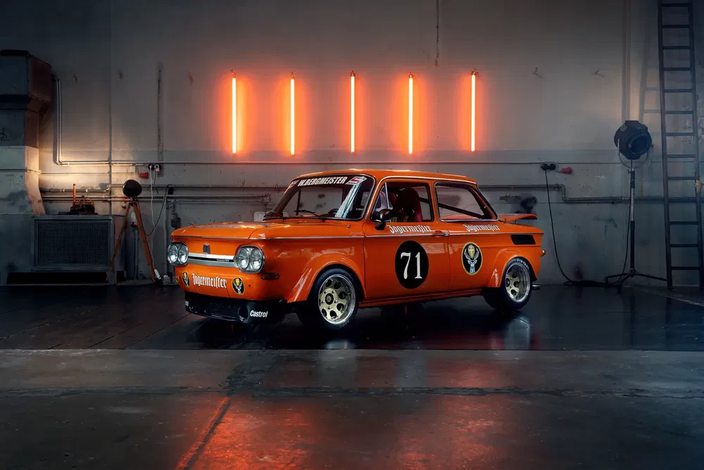 Retro oranje NSU raceauto met nummer 71 in een industriële vintage garageomgeving.