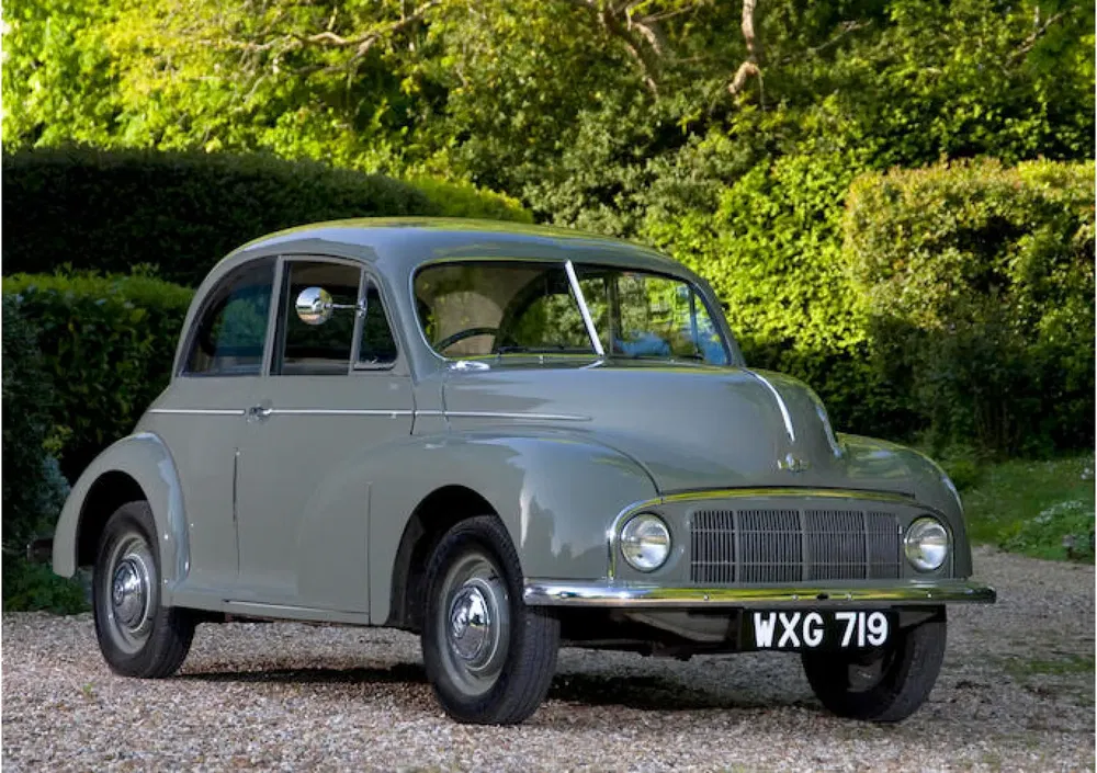 Voiture Morris Minor grise stationnée sur une allée de gravier, entourée de verdure.
