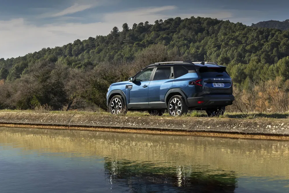 Blauwe Dacia SUV geparkeerd nabij een meer, bos op de achtergrond.
