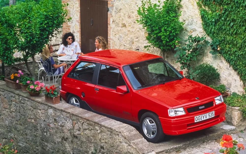 Opel Corsa rouge garée près d'une terrasse en pierre avec des plantes autour.