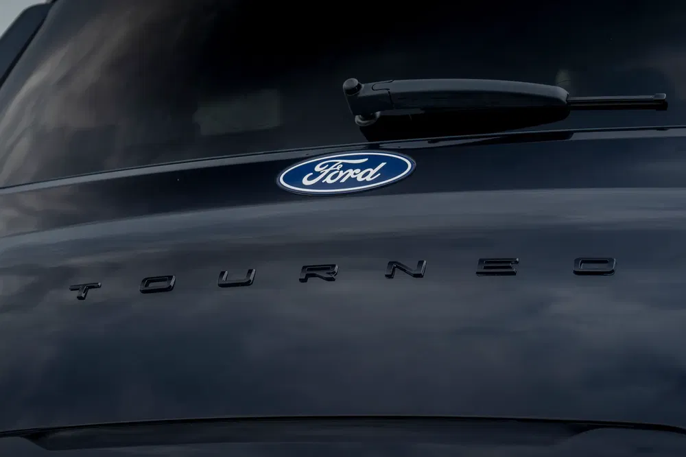 Logo Ford sur hayon Ford Tourneo, emblème et modèle en arrière-plan sombre.