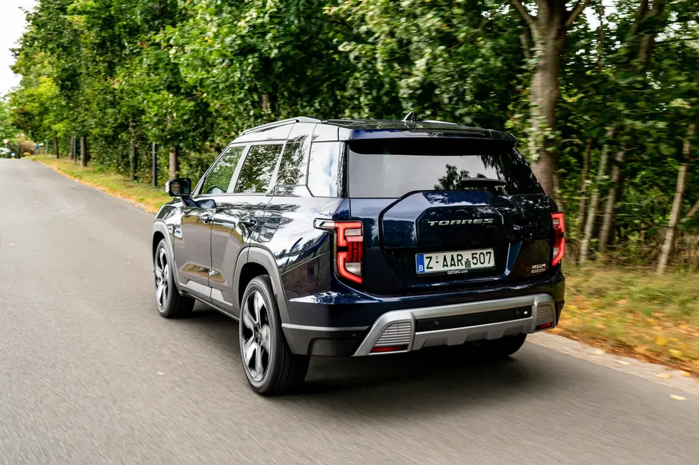 SUV SsangYong Torres roulant sur route arborée, vue arrière en mouvement.