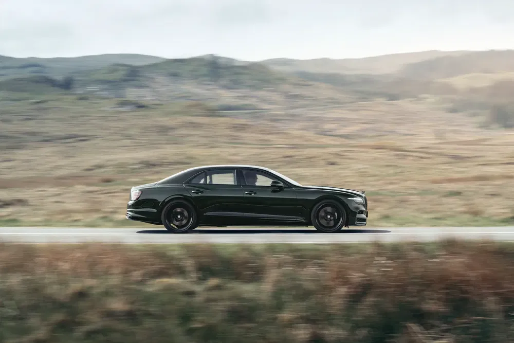 Bentley Flying Spur roulant sur une route panoramique, paysages vallonnés en arrière-plan.