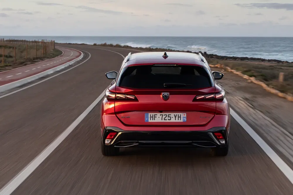 Rode Peugeot 308 op kronkelende kustweg, schilderachtige oceaan op de achtergrond.
