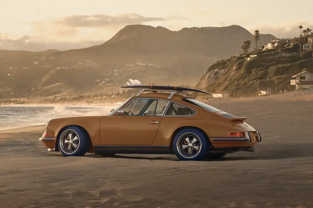 Porsche classique sur plage avec planche de surf, paysage côtier et collines en arrière-plan.