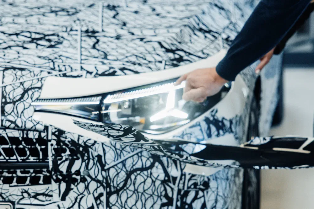 {"fr":"Prototype de voiture camouflée, feux avant design futuriste, main inspectant la carrosserie.","nl":"Geïncamoufleerde auto-prototype, futuristisch ontworpen koplampen, hand inspecteert carrosserie."}