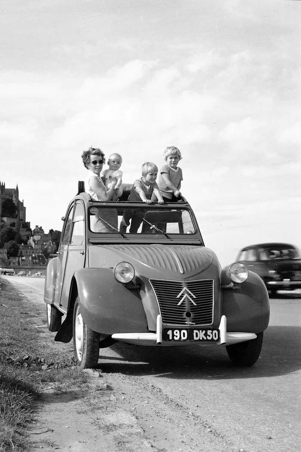 Citroën 2CV avec femme et enfants profitant du paysage côtier en arrière-plan.