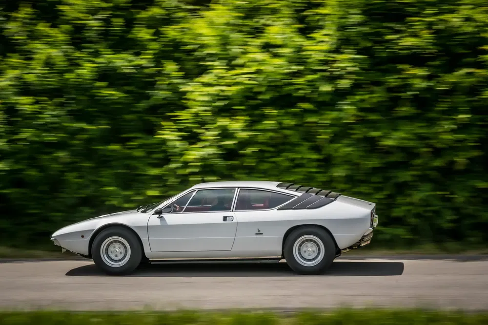 Zilveren Lamborghini Jarama rijdend op een weg omgeven door groen.