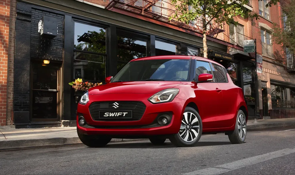 Rode Suzuki Swift geparkeerd voor een bakstenen gebouw in een levendige straat.